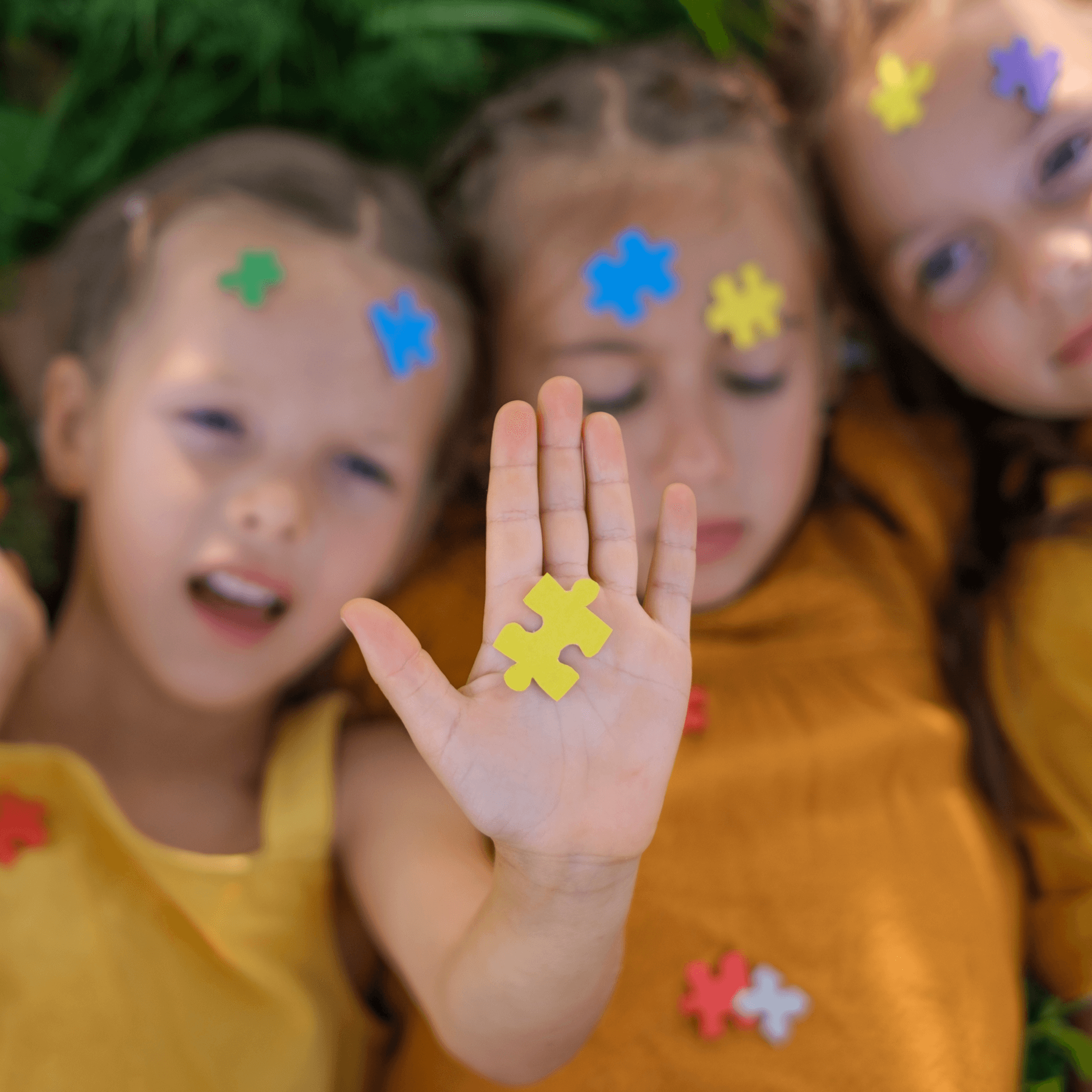 Three young girls laying in grass with puzzle pieces on faces and body. One girl is holding up the palm of her hand, with a puzzle yellow puzzle piece in the centre.