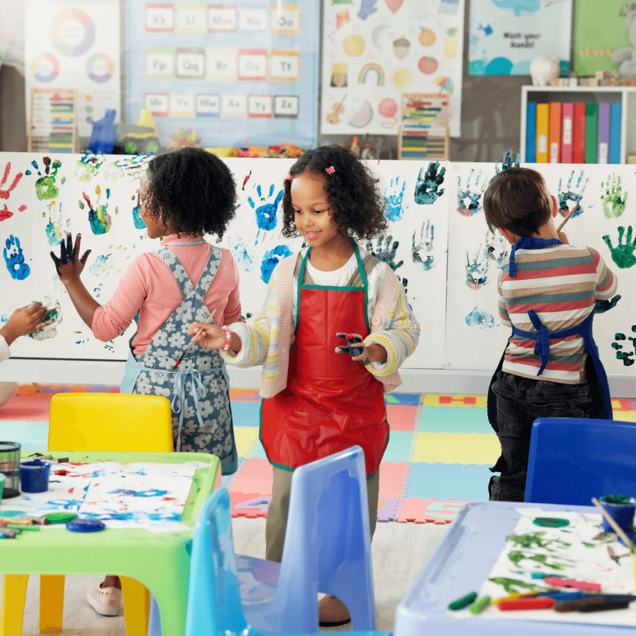 Group of children in a classroom painting together. Canvases with painted handprints in the background.