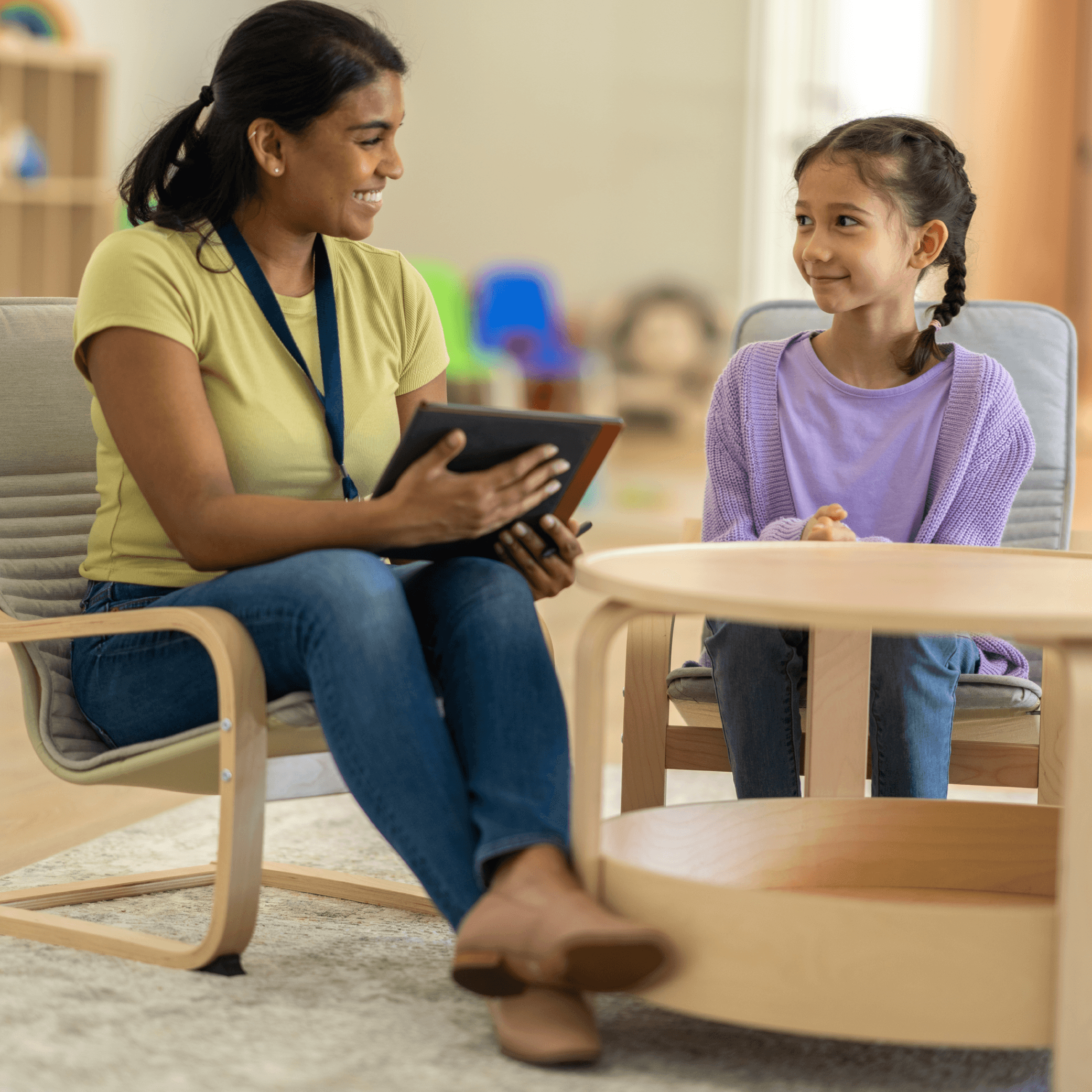 Young girl sitting in chair beside a woman in a counselling-like setting. Both are smiling at each other. Woman is holding a tablet.