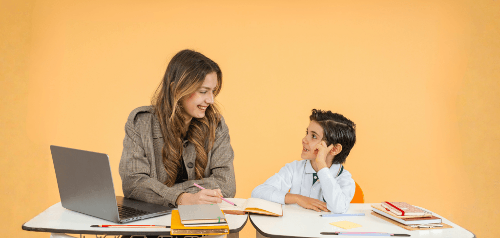 Female teacher and young boy sitting beside each other at work desks, looking at one another smiling.